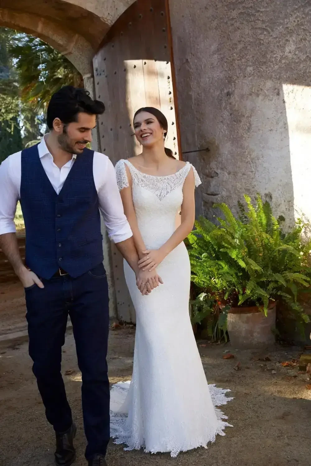 A couple walking hand in hand. The woman is wearing a white wedding dress, and the man is dressed in a blue vest with a white shirt and dark pants. They are standing near an old stone structure with greenery around them.