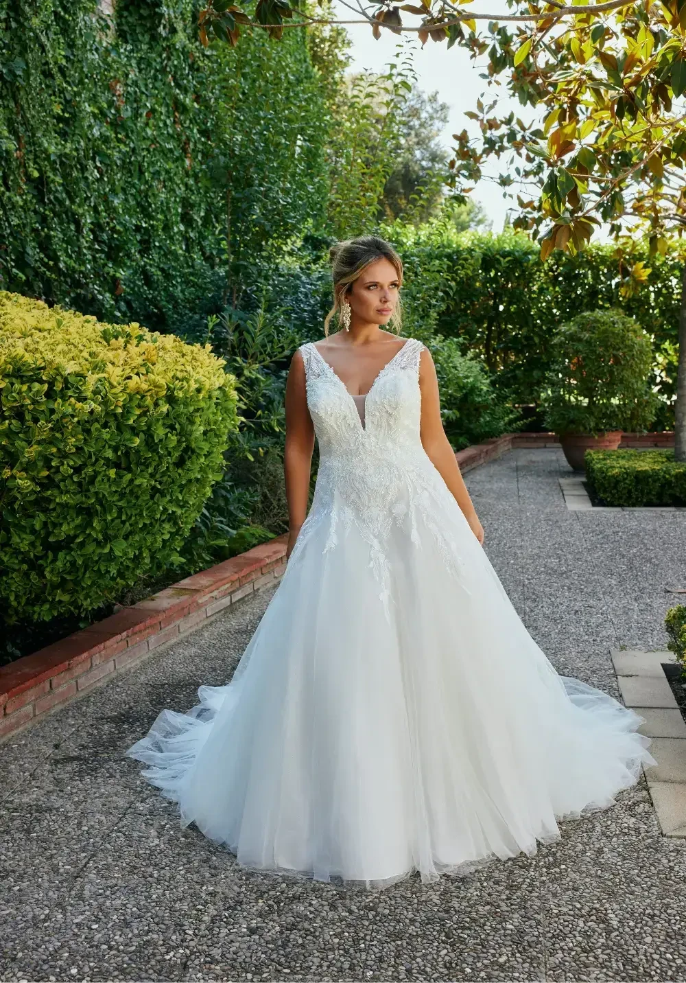 A woman wearing a white wedding dress stands elegantly in a garden, surrounded by greenery.