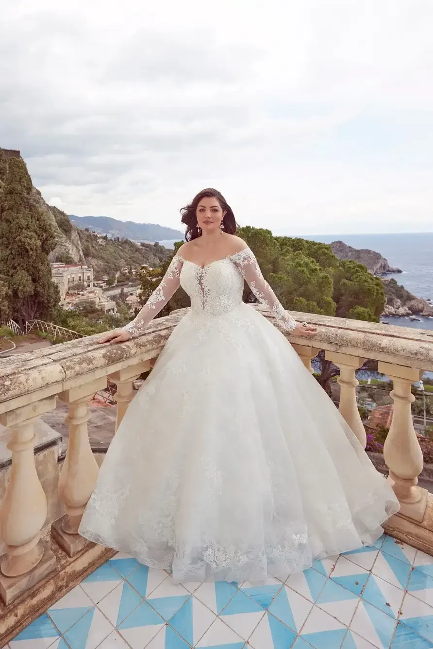 A woman in a beautiful white ball gown with long lace sleeves stands on a balcony overlooking a coastal landscape. The dress has intricate detailing and the background features hills and ocean views.