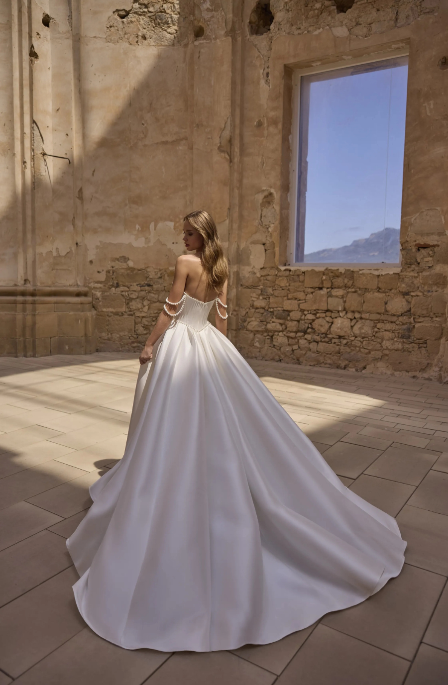 A woman in a flowing white wedding gown stands with her back to the camera, inside a rustic room with stone walls and a window, capturing soft sunlight.