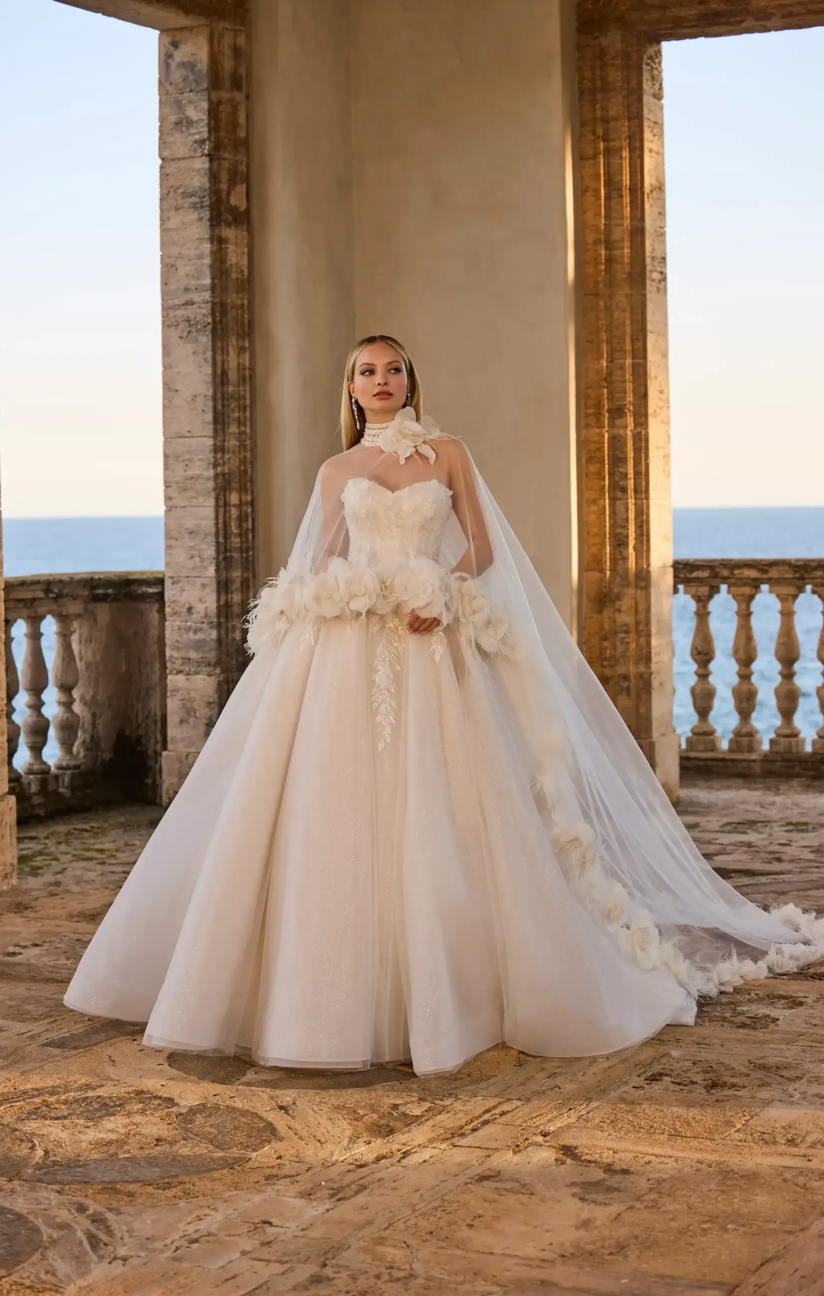 A bride wearing a strapless wedding gown with floral details and a flowing veil, standing in an elegant outdoor setting with a seaside view.