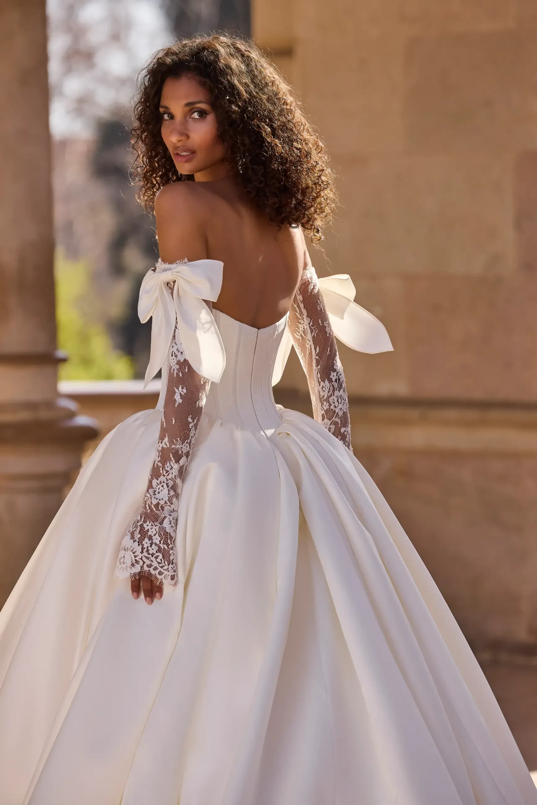Bride in elegant, off-shoulder gown with lace sleeves and large bows, stands in a sunlit stone courtyard. She gazes over her shoulder, exuding grace.