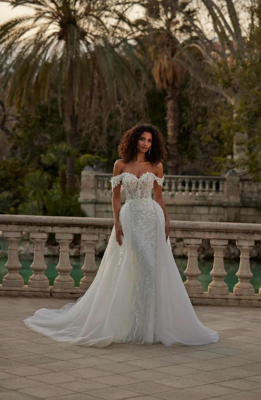 A woman in an elegant off-the-shoulder wedding dress stands on a stone terrace surrounded by palm trees.