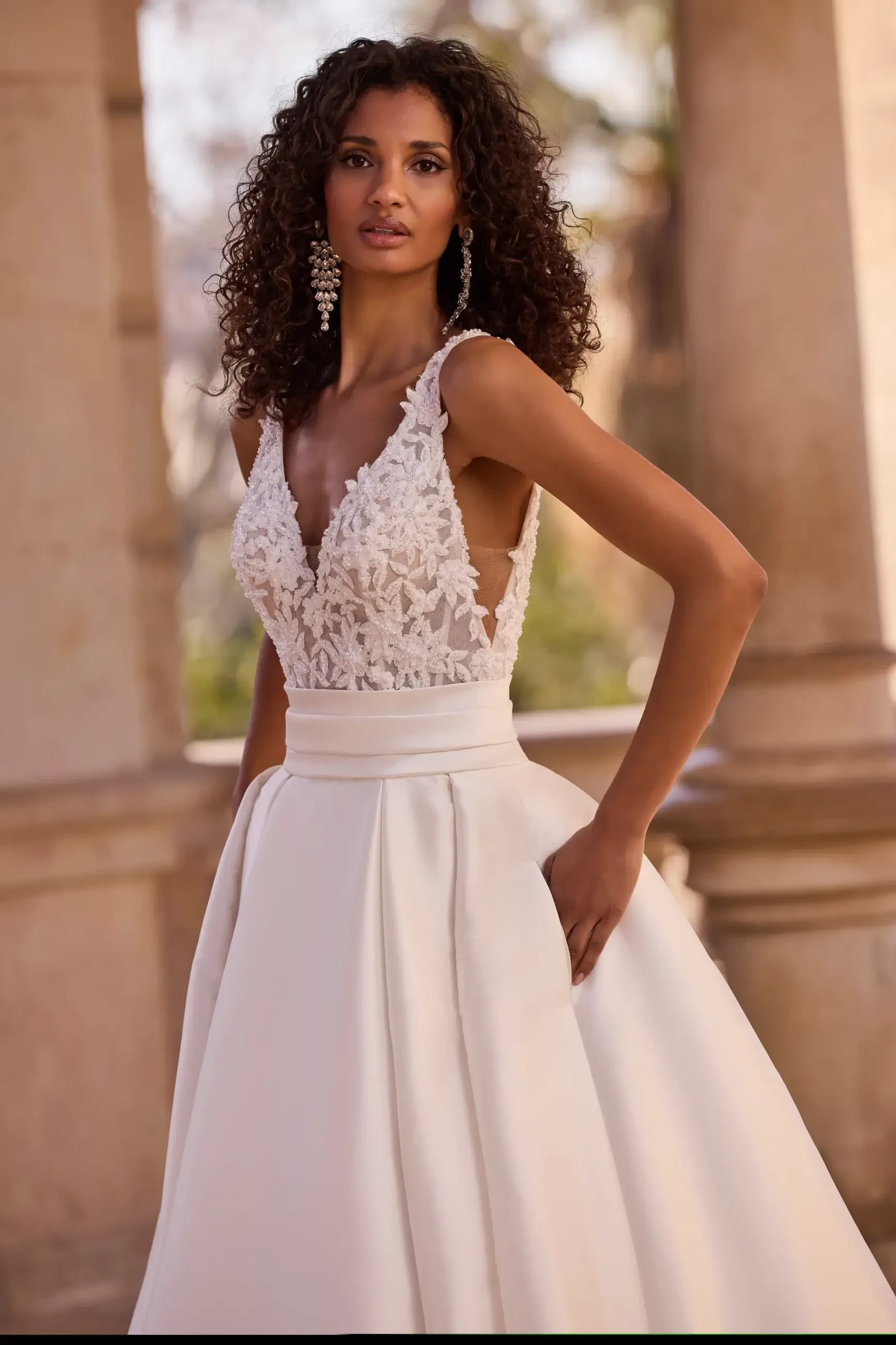 Woman in an elegant, sleeveless white lace wedding dress with a full skirt, standing confidently against a blurred outdoor background.