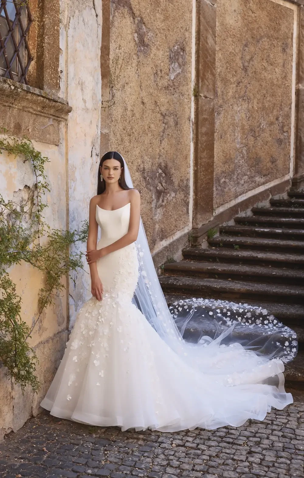 A bride in a white wedding gown with floral embellishments stands on a staircase near a textured wall, holding her veil.