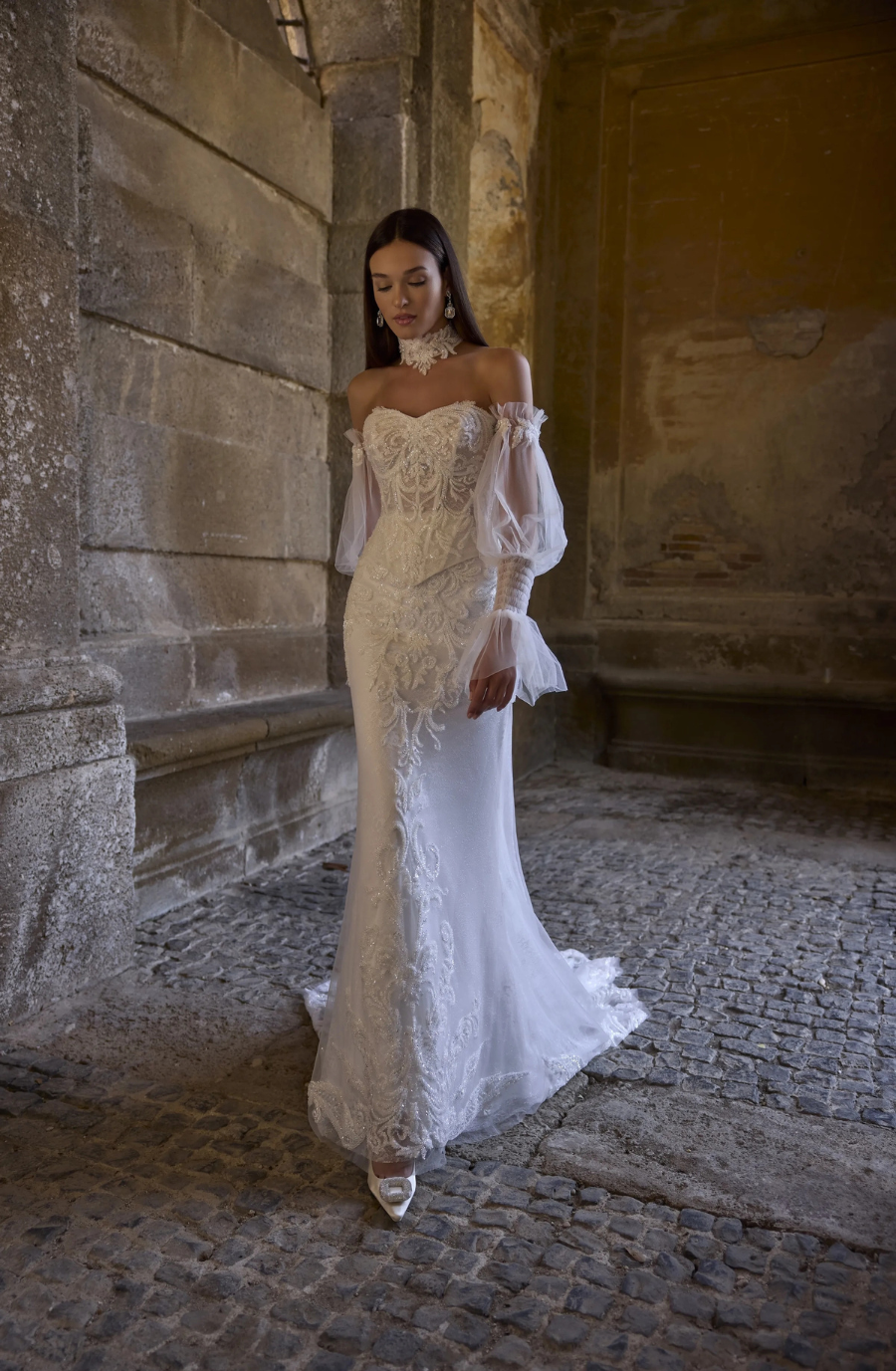 A woman wearing an elegant white off-shoulder lace gown with sheer sleeves, walking in a stone-walled interior.