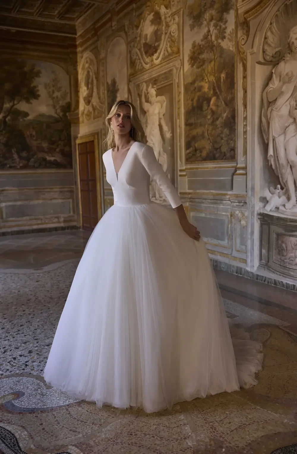 A woman in a white wedding dress standing in an ornate room with classical sculptures and paintings.