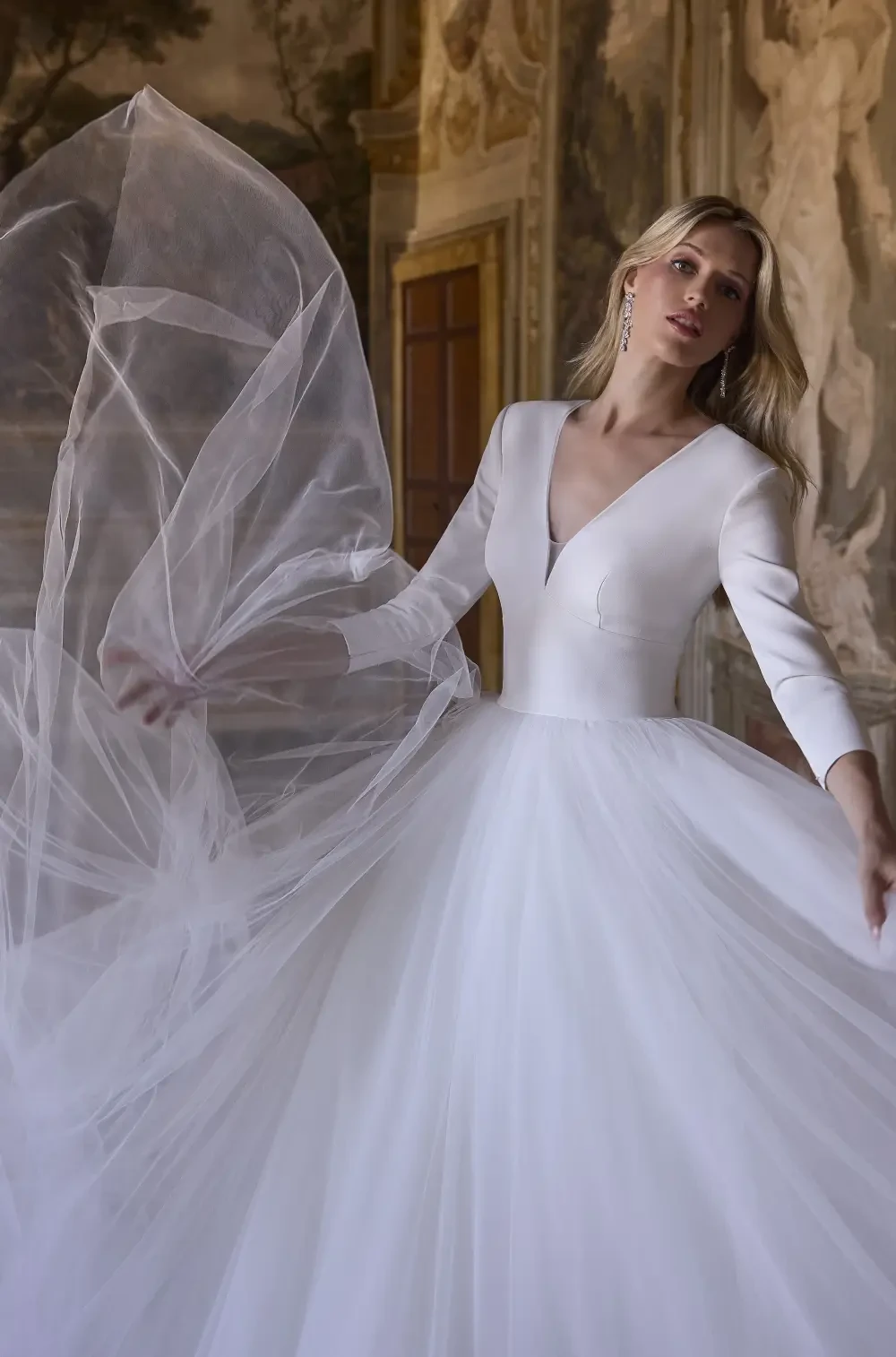 A woman in a white wedding dress standing in an ornate room with classical sculptures and paintings.