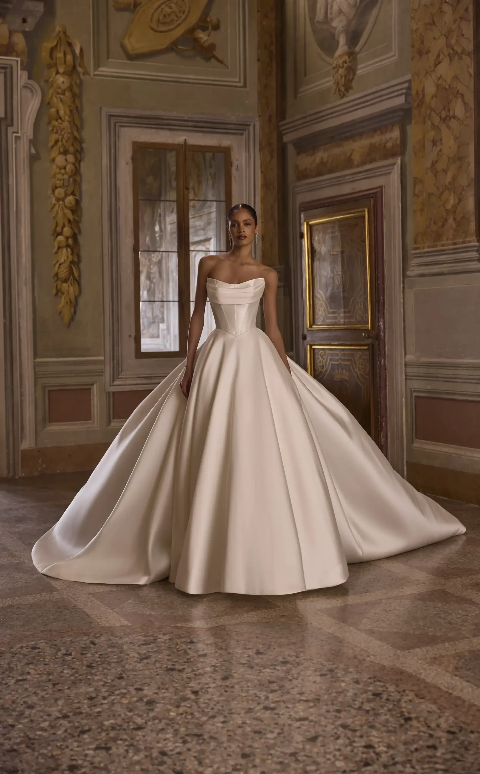 A model wearing a strapless, white ball gown in an elegant room with ornate decorations.