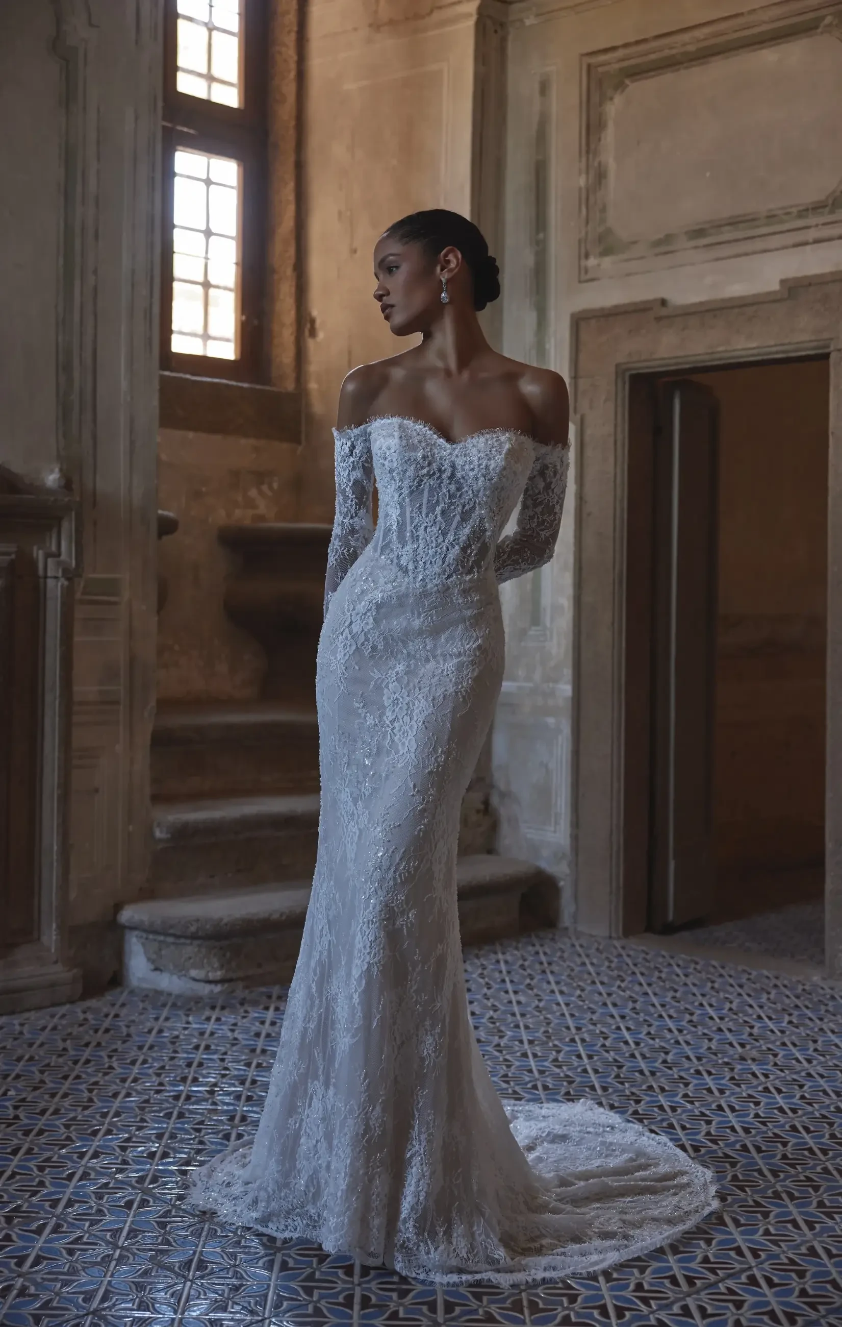 A model in a fitted, off-the-shoulder white lace gown stands elegantly in a historic interior with wooden windows and patterned tile flooring.