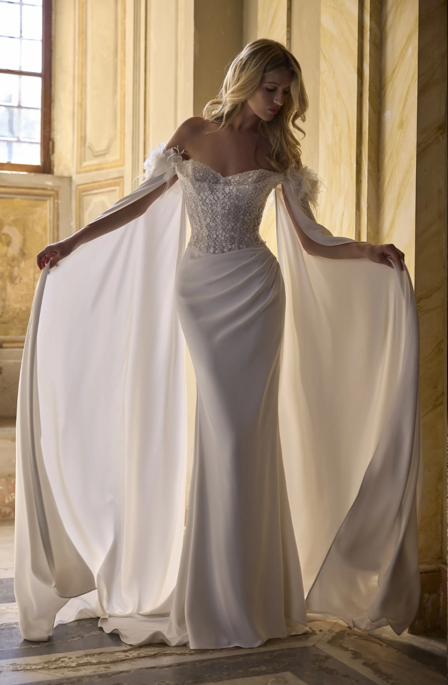 A woman in a flowing, elegant white gown with off-the-shoulder sleeves and a cape-like train, posing in an ornate interior setting.