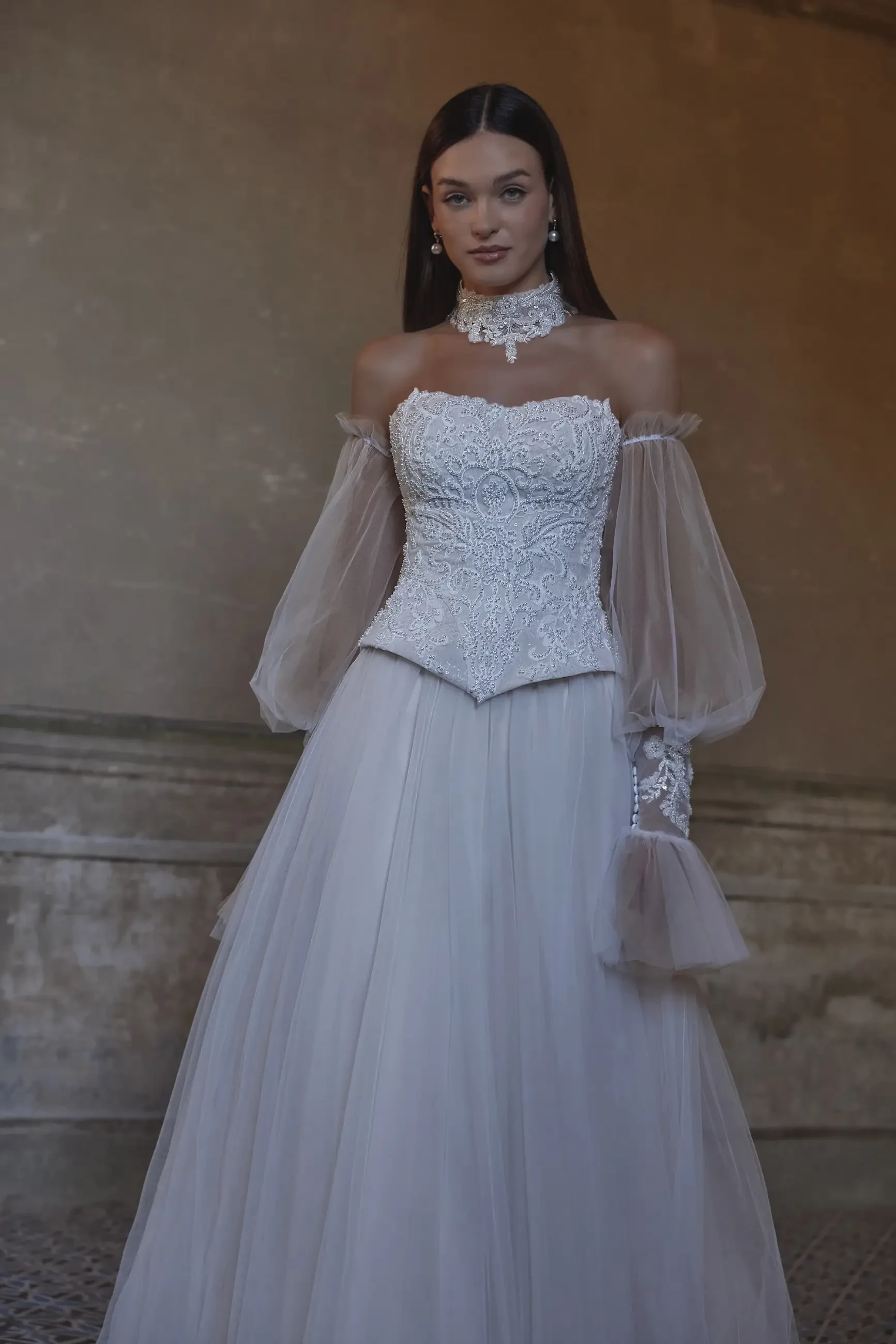 A model wearing a white wedding dress with a fitted bodice and sheer puff sleeves, posing in an elegant setting.