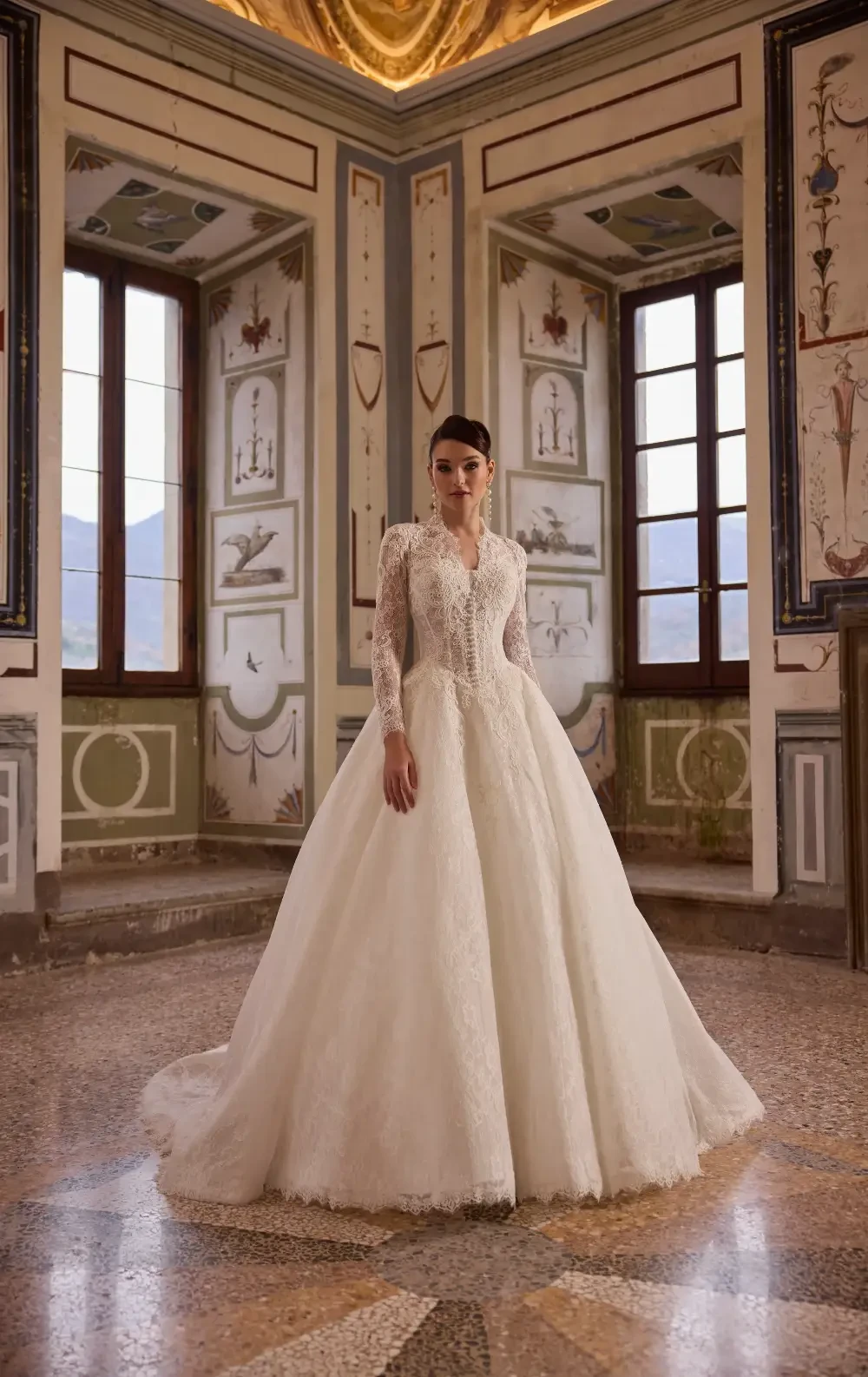A bride in a luxurious white gown stands elegantly in a beautifully decorated room with large windows and ornate walls.