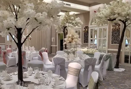 Elegant wedding venue with white floral centerpieces and trees, white draped chairs, and mirrored walls. Soft, romantic ambiance with muted tones.